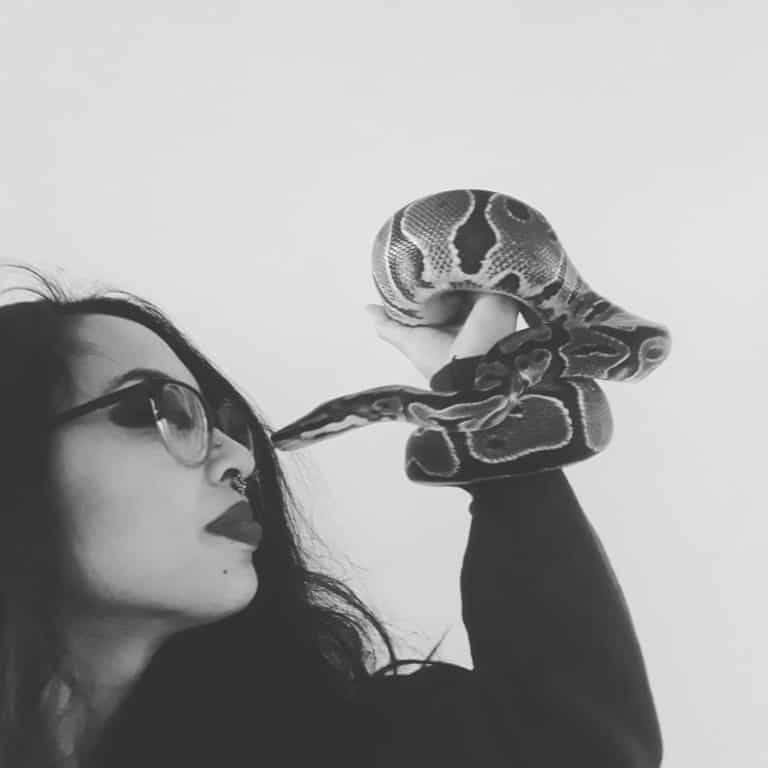 Profile of nonbinary light brown skinned astrologer with thick glasses, holding ball python; their noses touch